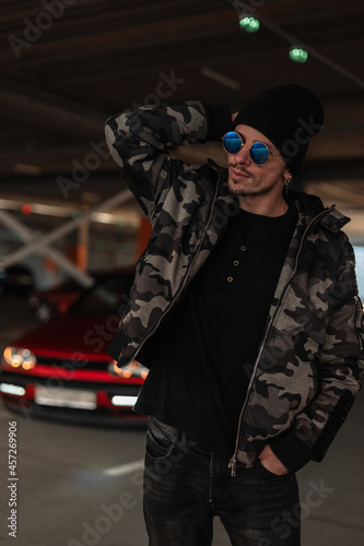 fashion hipster man with blue sunglasses in winter military jacket and pullover with hat near red car in city