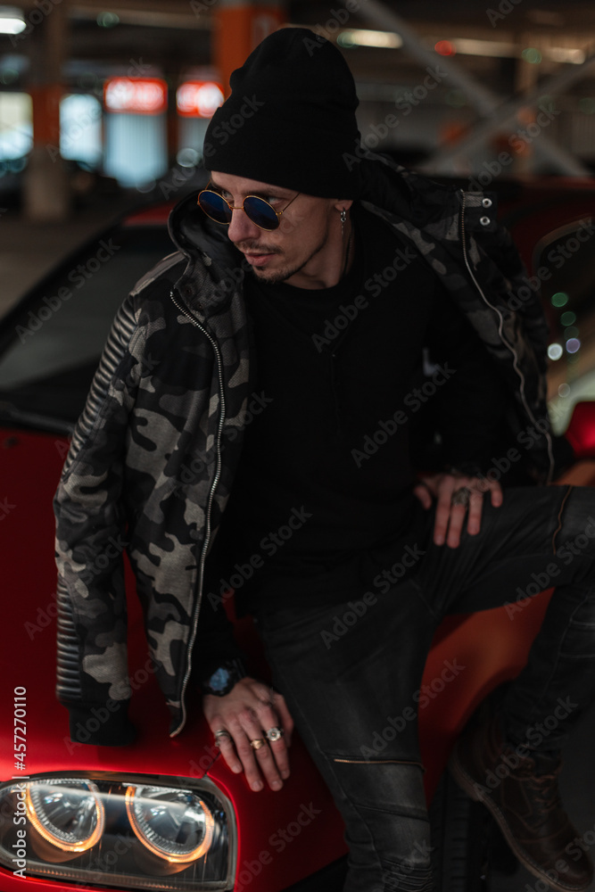 Stylish cool handsome model man with vintage sunglasses in a fashionable military jacket with a black hat and pullover sits near a red car on the street