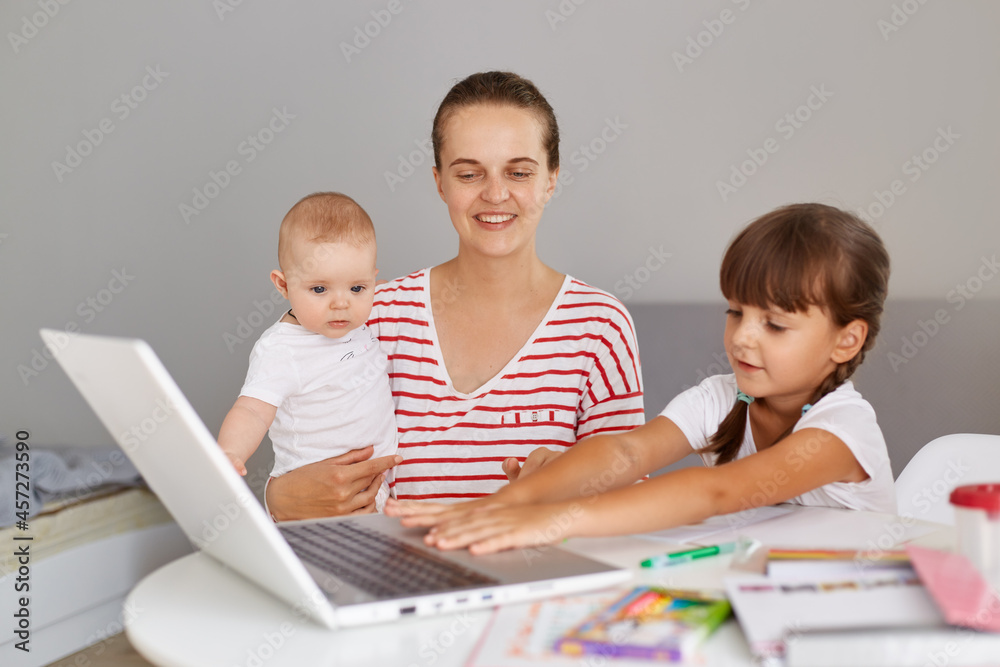 Happy positive young adult mother with baby in hands sitting at table and helps her elder daughter to do home task or helping during lesson, looking smiling at laptop computer. Happy positive young adult mother with baby in hands sitting at table and helps her elder daughter to do home task or helping during lesson, looking smiling at laptop computer.