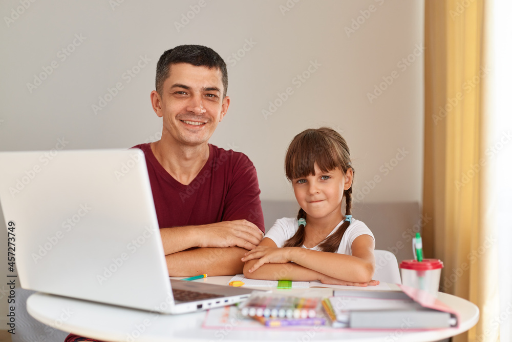 Indoor shot of smiling handsome man sitting at table with his little daughter in front of laptop, looking at camera with happy expression, distant education. Indoor shot of smiling handsome man sitting at table with his little daughter in front of laptop, looking at camera with happy expression, distant education.