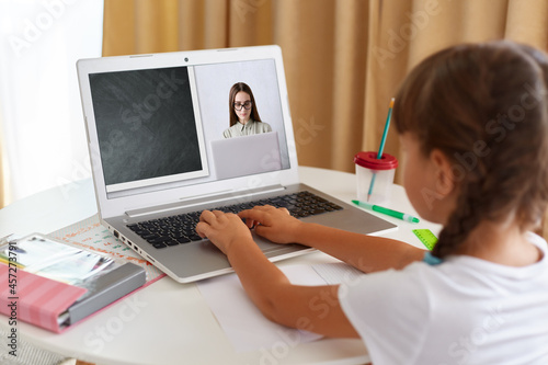 Back view portrait of schoolgirl wearing white t shirt sitting in front of laptop and looking at screen, having online lesson via web camera, distant education.