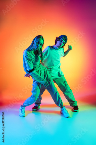 Young dancers, stylish emotive girl and boy dancing hip-hop in casual sports youth clothes on gradient multi colored background at dance hall in neon light.