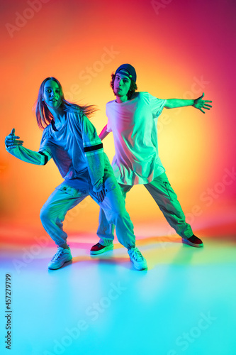 Young hip-hop dancers, stylish emotive girl and boy in action and motion in casual sports youth clothes on gradient multi colored background at dance hall in neon light.