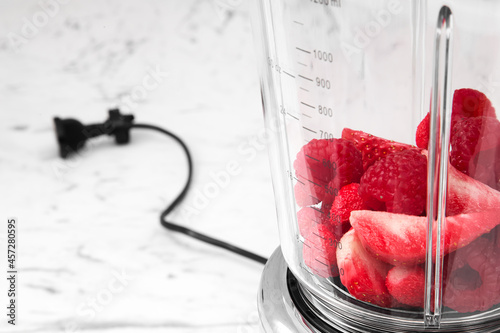A blender with raspberries and strawberries stands on a white countertop in the kitchen. Horizontal theme for healthy lifestyle and nutrition