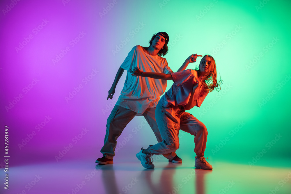 Stylish young hip-hop dancers, emotive girl and boy in action and motion in casual sports youth clothes on gradient multi colored background at dance hall in neon light.