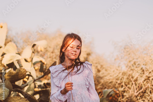 Thoughtful preteen girl wearing eco style boho dress and walking alone in summer field near big desert cactus.
