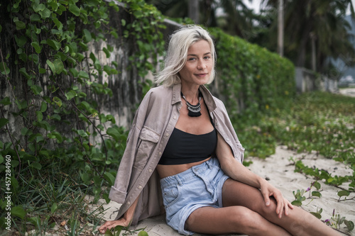 Portrait of pretty caucasian young woman with blond hair, wearing casual clothing, sitting on the beach with grass and enjoying 