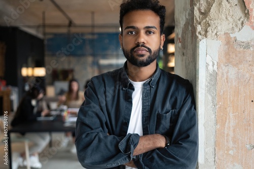 Portrait of mixed-race young man standing in office with colleagues meeting in background