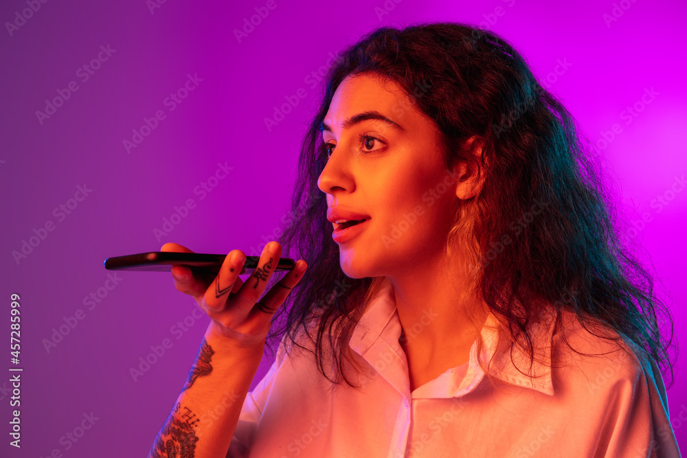 Fototapeta premium Bright fashionable portrait of beautiful happy girl using phone isolated on purple, lilac color studio background in neon light.