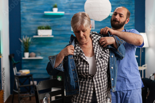 Social asisstant man worker helping pensioner disabled senior woman puts jacket during recovery session. Healthcare assistance. Social services nursing elderly retired woman. Disability people support