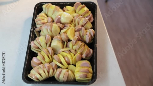 Sliced potatoes with sausages on a baking sheet are sprinkled with spices.