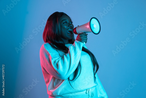 One African beautiful woman with long straight hair shouting at megaphone isolated on blue studio background in neon light. Concept of human emotions