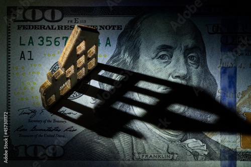 A small conventional wooden house stands on a fragment of a 100 American dollar bill and casts a long shadow over the area of ​​the bill.