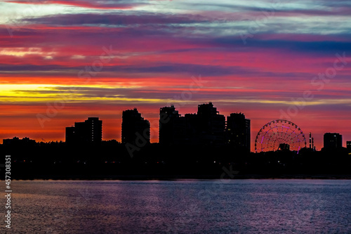 Picturesque sky above silhouettes of houses of modern city. Buildings are located on coastline. Ferris wheel is also visible
