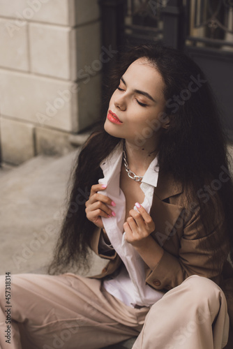 Close up portrait of young beautiful woman with long brunette curly hair posing against building background. sitting on the stairs