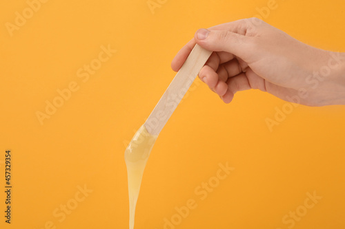 Woman holding spatula with hot depilatory wax on yellow background, closeup