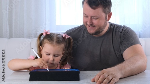 little girl 3 years old and her father draw draws on the tablet and at home watching a drawing lesson online. Dad rejoices at his daughters success and helps her in her studies. Online school distance