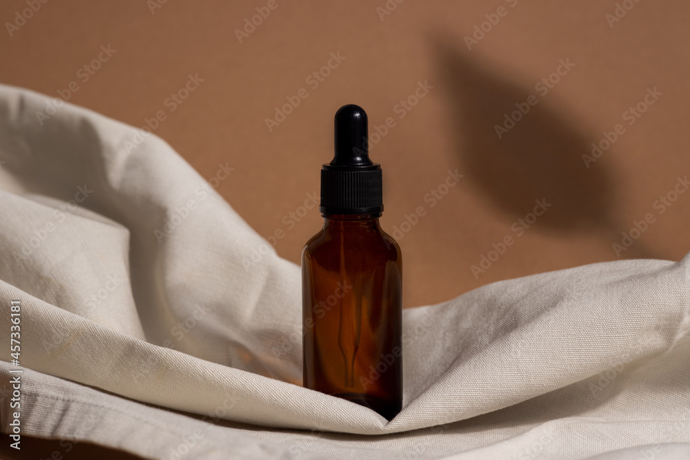 Creative photo of cosmetic bottle with pipette on beige fabric and brown background with shadow of tropical flower. Advertising