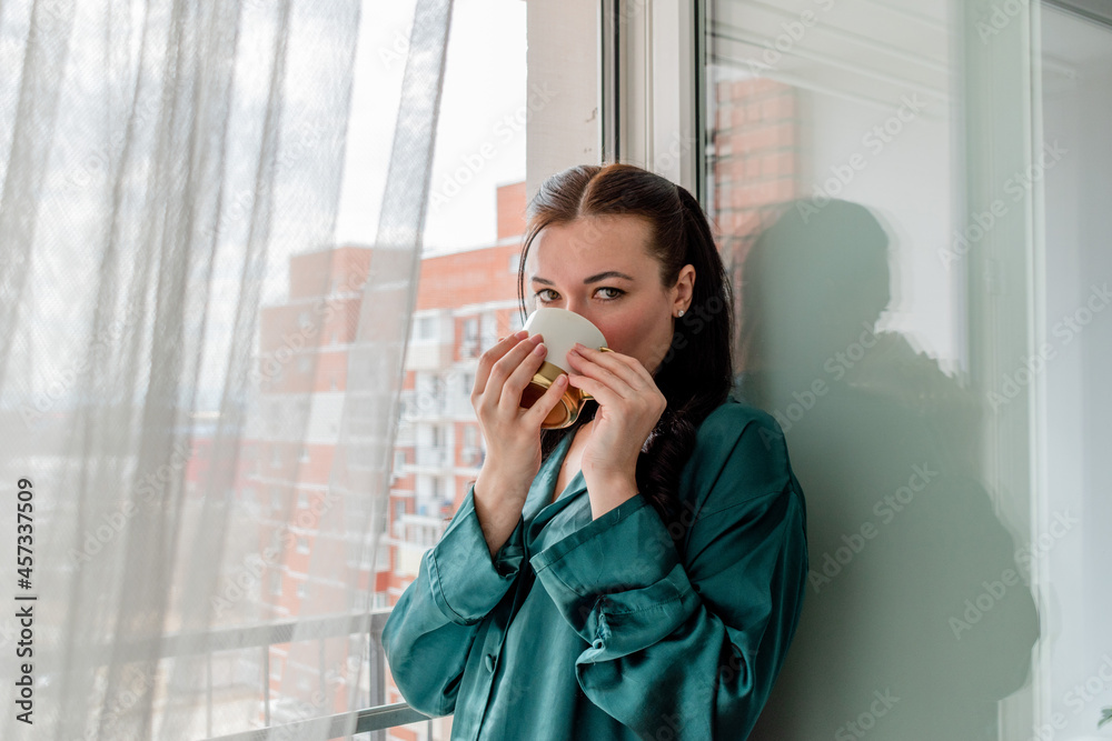 Young woman drinks coffee from mug while standing near the window. Home comfort zone and wellness concept. Young woman drinks coffee from mug while standing near the window. Home comfort zone and wellness concept.