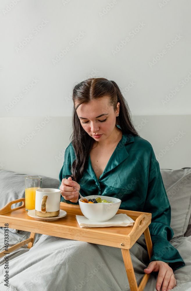 Young woman in silk pajamas eating her breakfast while lying in bed. Wellness concept.