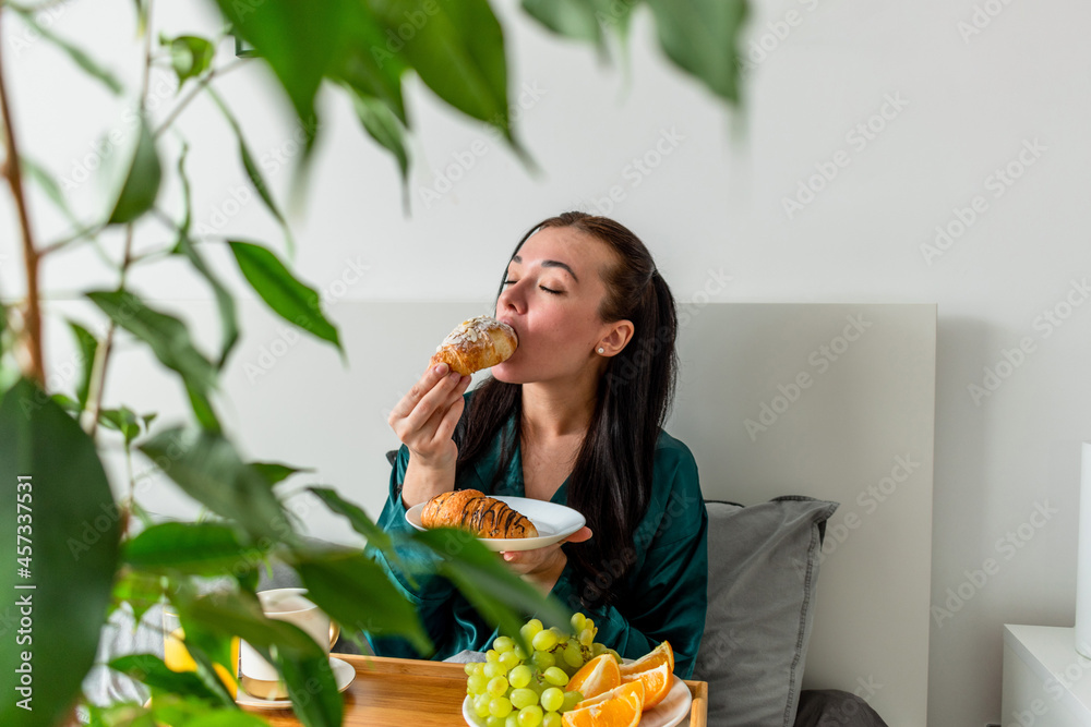 Young woman in silk pajamas has healthy breakfast while lying on bed in bedroom. Home wellbeing concept. Emotional health of a young woman Young woman in silk pajamas has healthy breakfast while lying on bed in bedroom. Home wellbeing concept. Emotional health of a young woman