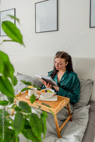 Young woman in silk pajamas has healthy breakfast and read book while lying on bed in bedroom.