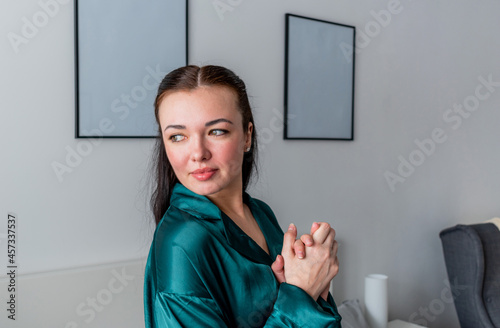 Young woman with dark hair silk pajamas looks out the window. Wellness concept
