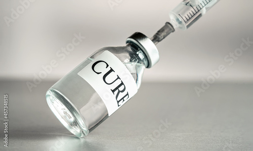 Glass vaccine vial with label Cure on white board, hypodermic syringe needle inside  - vaccination concept