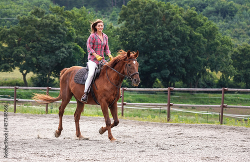 Young woman in shirt riding on brown horse, smiling, hair moving in air because of speed, blurred trees background