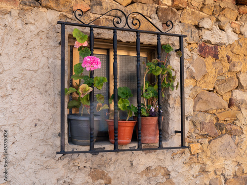 A closeup of a window with black bars with sliding glass in a wall of a stone and adobe house and decorated with geraniums in pots