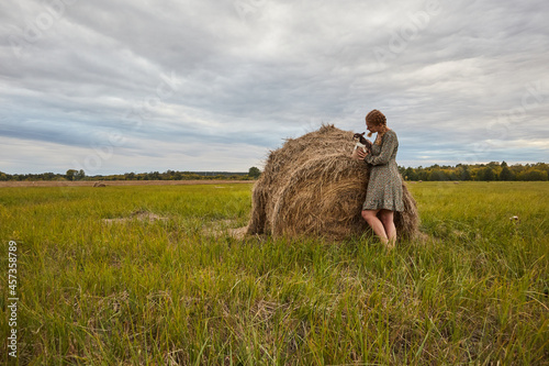 blonde girl in a dress with a basket and a dog standing in a field near a haystack