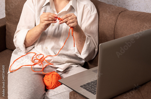 A woman learns to crochet clothes from knitted yarn, remotely via a laptop at home. Selective focus.