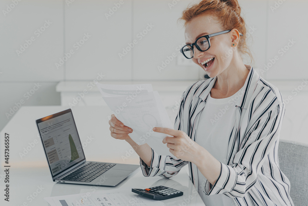 Overjoyed ginger woman reading reading financial paper document and smiling of pleasant news Overjoyed ginger woman reading reading financial paper document and smiling of pleasant news