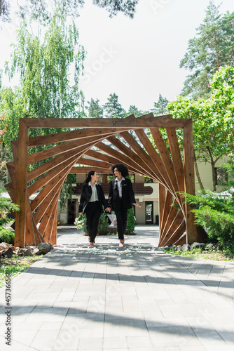 Smiling multiethnic lesbian couple in suits with wedding bouquet walking in park. © LIGHTFIELD STUDIOS