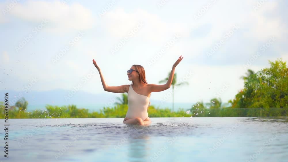 Happy Woman in One-shoulder Monokini and Sunglasses Splashing Water with Hands while Sits on the Edge of an Exotic Resort Pool with Feet in the Water, front slow-motion Happy Woman in One-shoulder Monokini and Sunglasses Splashing Water with Hands while Sits on the Edge of an Exotic Resort Pool with Feet in the Water, front slow-motion
