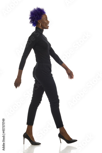 Confident African American Woman Is Walking In Black Clothes And High Heels. Full Length, Side View.