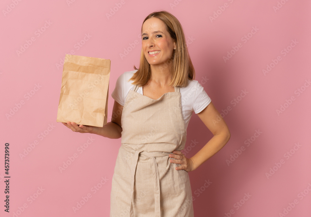 Smiling happy young woman in apron holding brown empty blank craft paper bag for takeaway mock up isolated on pink background Smiling happy young woman in apron holding brown empty blank craft paper bag for takeaway mock up isolated on pink background