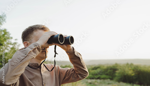 A young man looks through binoculars at a beautiful hilly landscape.The concept of hunting, travel and outdoor recreation. Banner with copy space.A traveler or hunter is observing through binoculars.