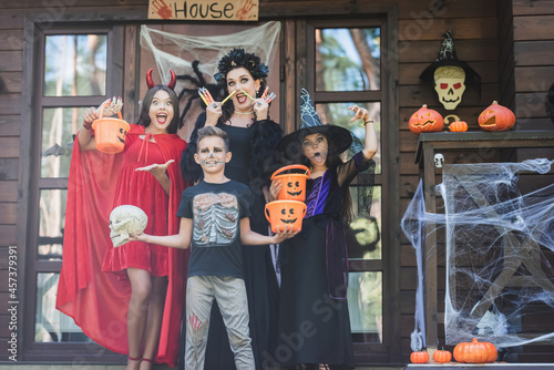 children with mom in halloween costumes standing with buckets, sweets and skull on porch with decoration