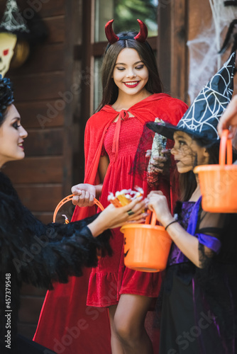 mom giving candies to happy daughters in devil and witch halloween costumes