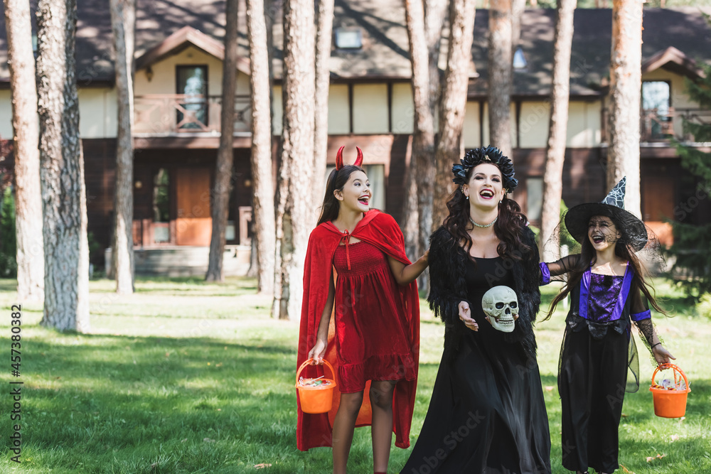 happy woman with daughters in halloween costumes laughing in forest near blurred cottage happy woman with daughters in halloween costumes laughing in forest near blurred cottage