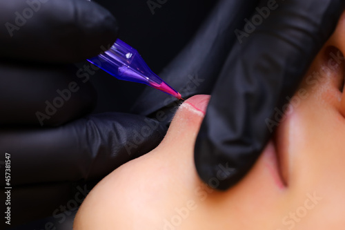 a tattoo artist in black gloves holds a tattoo machine  and the other finger presses on the client s lips  thereby performing a lip tattoo