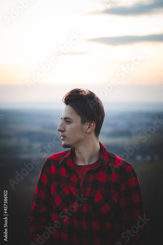 Candid portrait of a man in his 20s dressed in a black and red checked shirt looking into his future, immersed in his thoughts and contemplating. Authentic mood of a young man on an indecisive journey © Fauren
