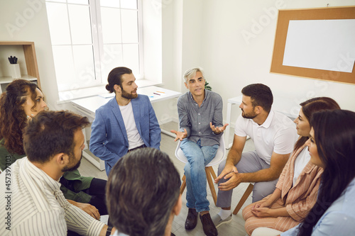 Group of people having serious conversation during work meeting in office. Senior man talking to team of employees or colleagues. Experienced coach sharing advice and providing business guidance