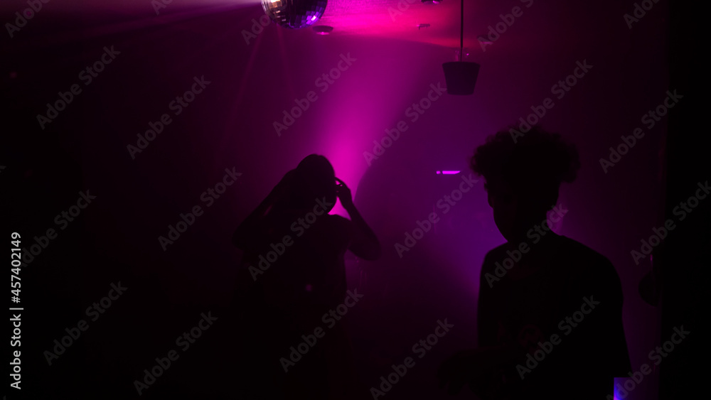 Silhouette image of people dance in disco night club to music from DJ on stage. Party atmosphere with disco ball. Nightlife concept. Space for text.