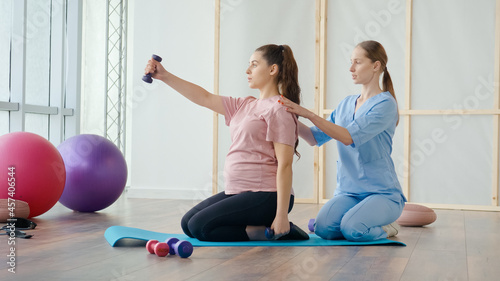 A Health Worker at a Medical Center Helps a Pregnant Woman to Do Exercises on a Ball with Dumbbells in Her Hands. The Expectant Mother Takes Care of her Health, Prepares for Childbirth.