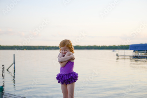 Little redhead girl in purple swimsuit hugging herself by a lake. photo