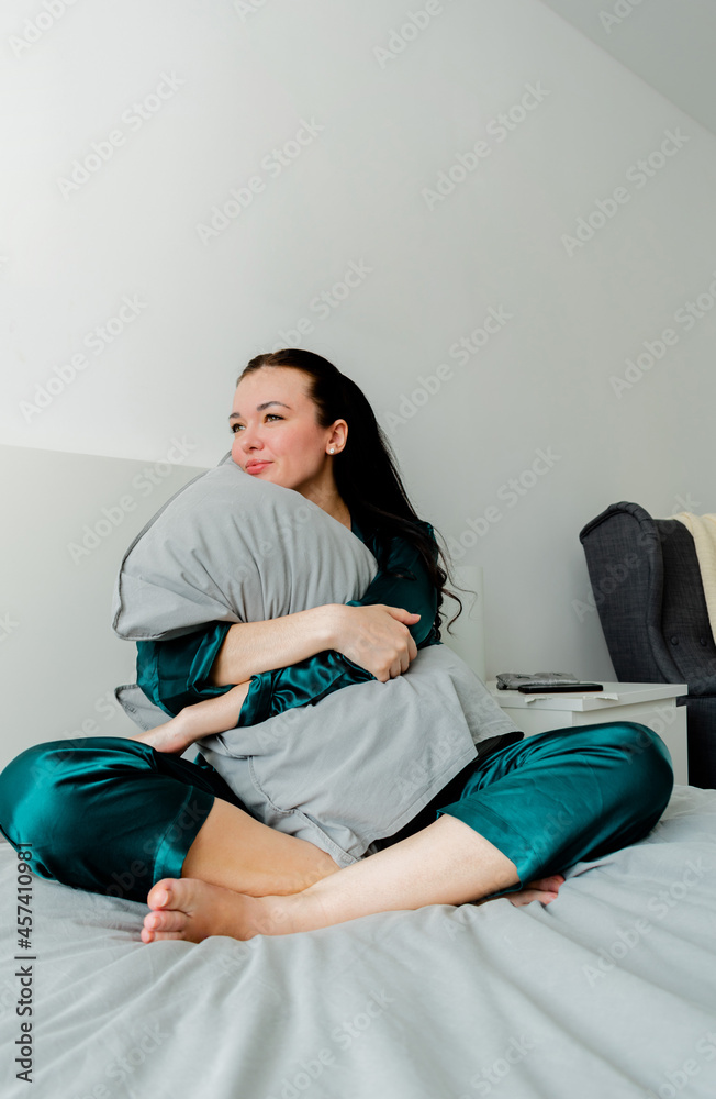 Obraz premium Young woman in silk pajamas sits on the bed and hugs a pillow. Loneliness inside the house. Wellness concept.