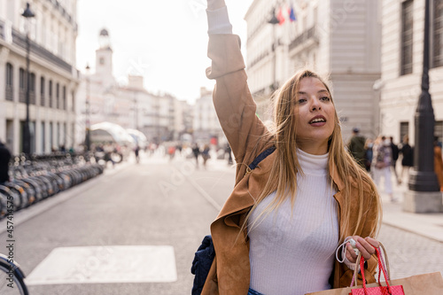 Smiling young female tourist waiting for cab outdoors. Cheerful modern lady with bag gesturing to give signal to taxi driver. Taxi service concept