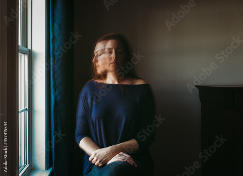 Woman with blurry face sitting alone in a dark room beside a window.